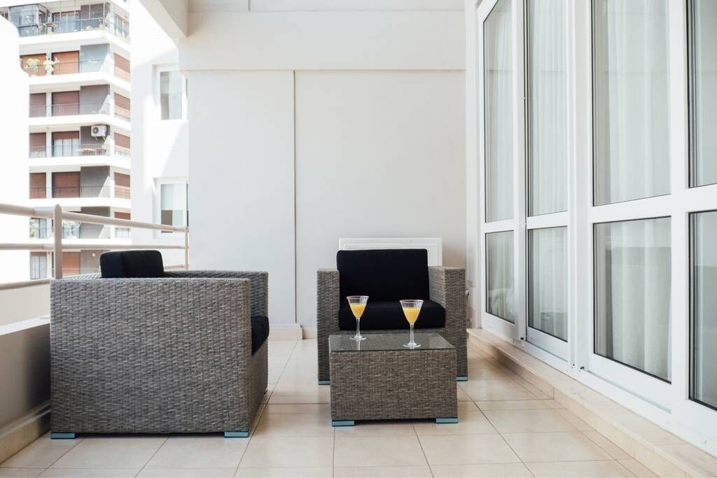A balcony with grey wicker furniture, two glasses of orange juice on a small table, and a view of a high-rise building.