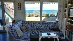 Ein Wohnzimmer mit einer Couch, einem Couchtisch und einem Bücherregal. Ein großes Fenster bietet Meerblick und Blick auf Grünflächen.