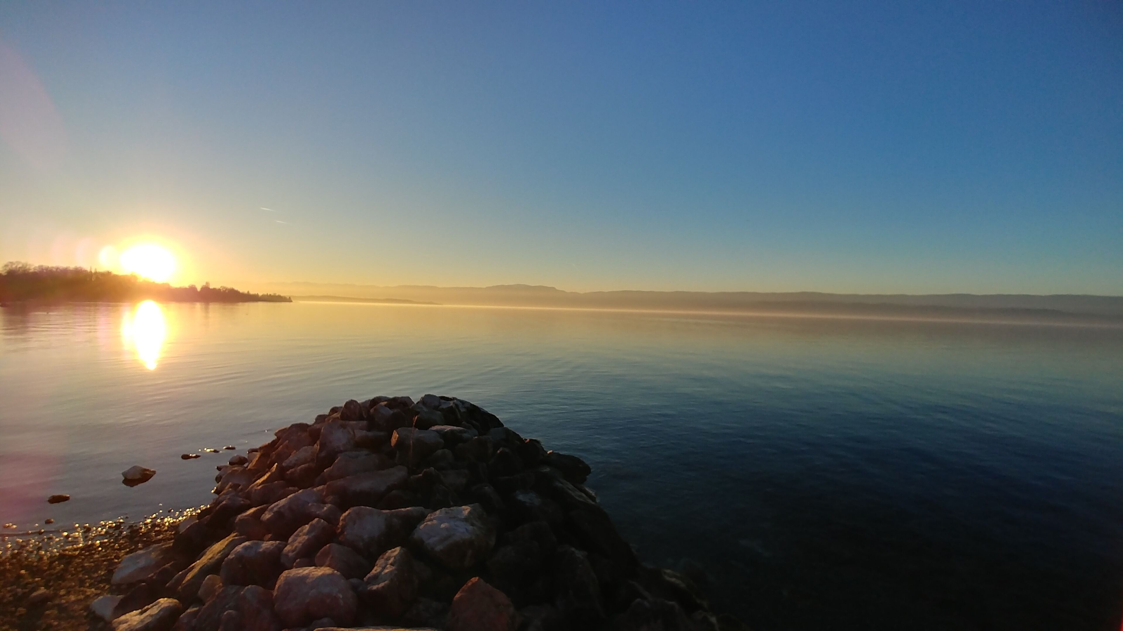 Ein ruhiger See bei Sonnenaufgang mit felsiger Uferlinie und Bergen im Hintergrund.