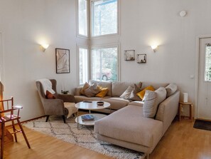 A modern living room with a sofa, armchairs, a coffee table, and a rug. There are two windows with blinds, wall-mounted lights, and framed pictures.