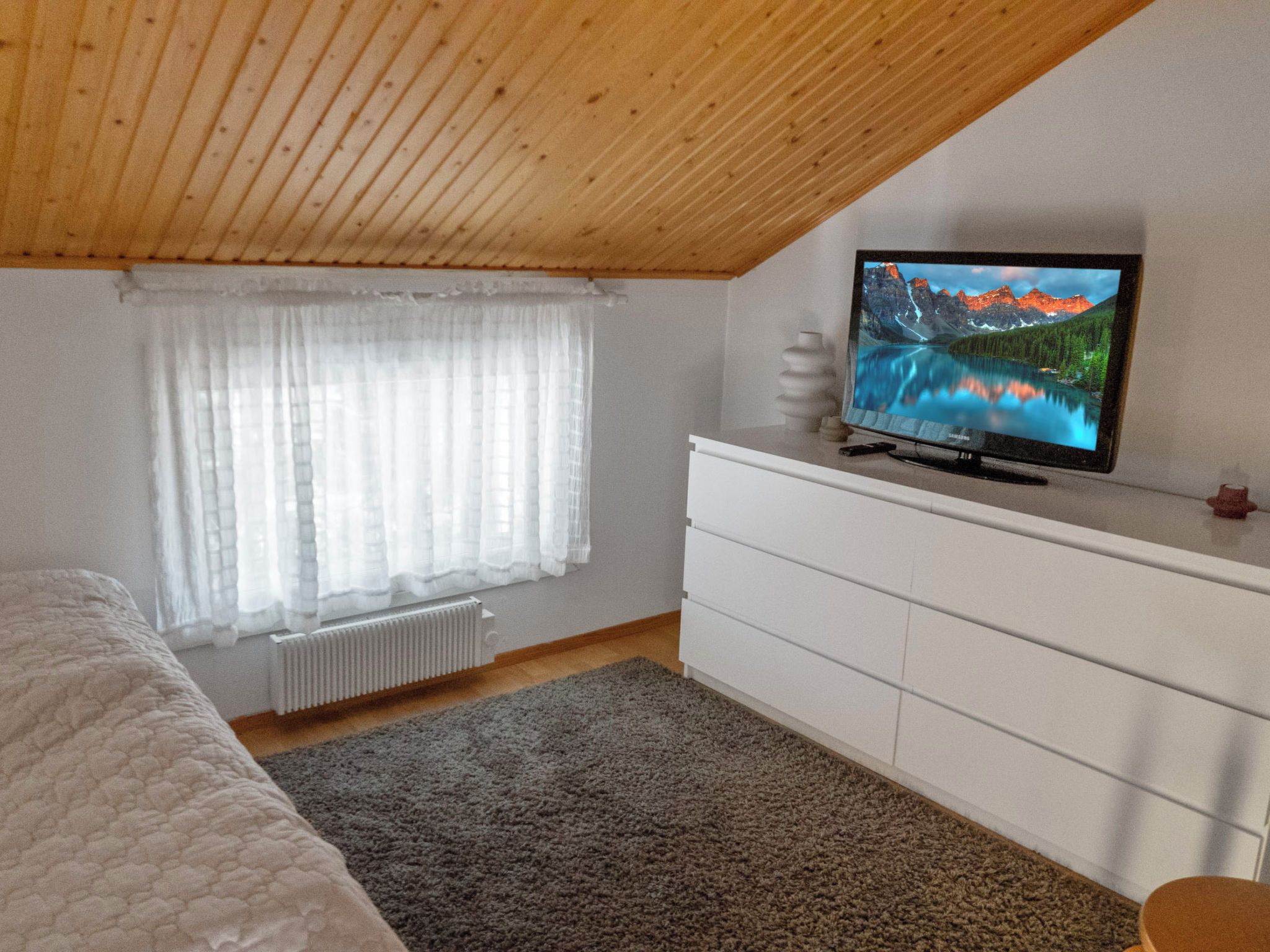 Ein Schlafzimmer mit Holzboden, einem Bett, einer weißen Kommode, einem Fernseher auf der Kommode und einem Fenster mit durchsichtigen Vorhängen.