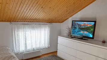 Ein Schlafzimmer mit Holzboden, einem Bett, einer weißen Kommode, einem Fernseher auf der Kommode und einem Fenster mit durchsichtigen Vorhängen.