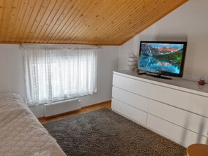 Quarto com teto de madeira, uma cama, cômoda branca, televisão sobre a cômoda e janela com cortinas finas.