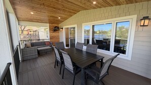 A covered patio with a wooden ceiling, a dining table with chairs, a sofa, and a view of a lake.