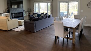 A modern living room with a fireplace, a grey sofa, a dining table with chairs, and large windows.