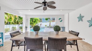Un comedor con mesa de madera y sillas de mimbre, ventilador de techo y vistas a una piscina y follaje exterior.