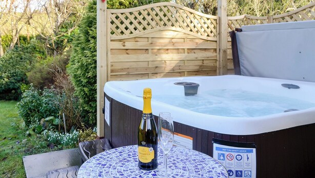 A hot tub with a bottle of Prosecco and a glass, surrounded by a wooden pergola and a garden.