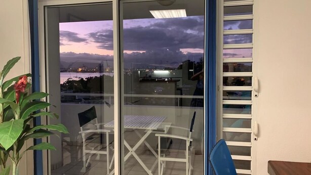 A balcony with a table and chairs, a potted plant, and a view of the cityscape at dusk. - Bleu Horizon Résidence by weekome (Les Abymes)