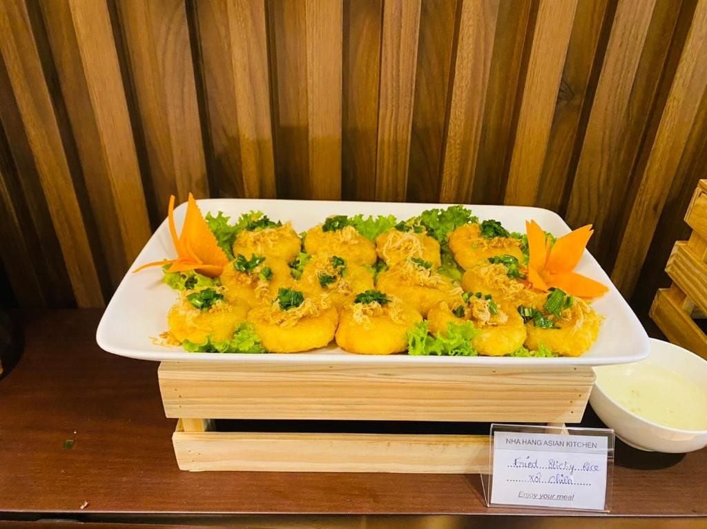 A rectangular white plate with yellow fried items, garnished with green herbs, on a wooden table with a sign reading 'NHA HANG ASIAN KITCHEN.'
