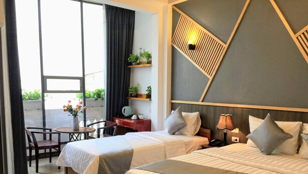 Ein Hotelzimmer mit zwei Betten, einem Schreibtisch mit Stuhl, einem kleinen Tisch mit Blumenvase und Blick auf Grünfläche.