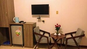 Ein gemütliches Zimmer mit Küchenzeile, einem wandmontierten Flachbildfernseher, zwei Sesseln, einem kleinen runden Tisch und einer Vase mit roten Blumen.
