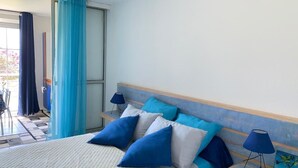 Cama bem arrumada com roupa de cama azul e branca, duas luminárias de cabeceira e vista para a varanda.