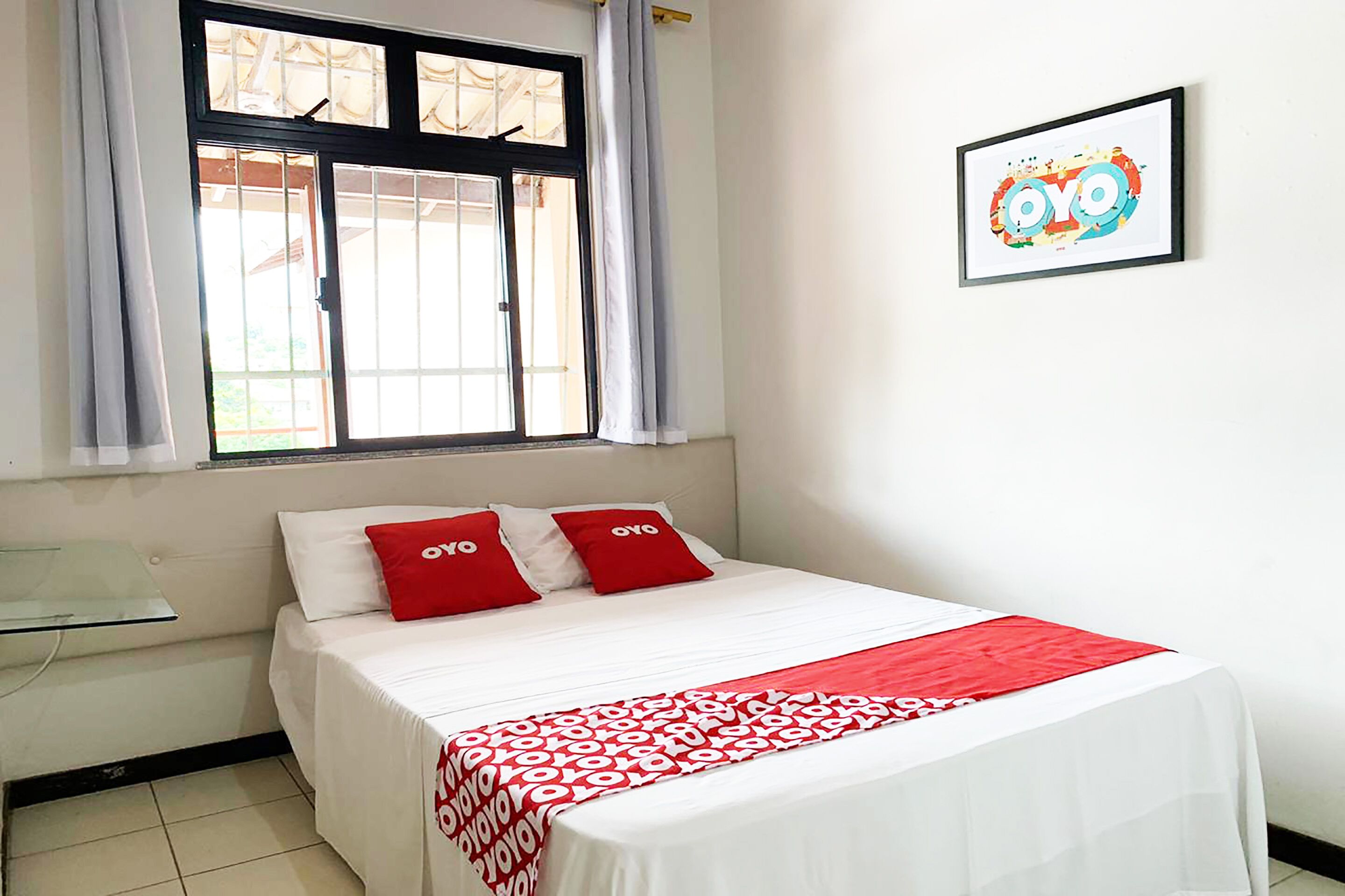 Una habitación de hotel con una cama, una ventana con cortinas, una mesita de noche y un cuadro en la pared.
