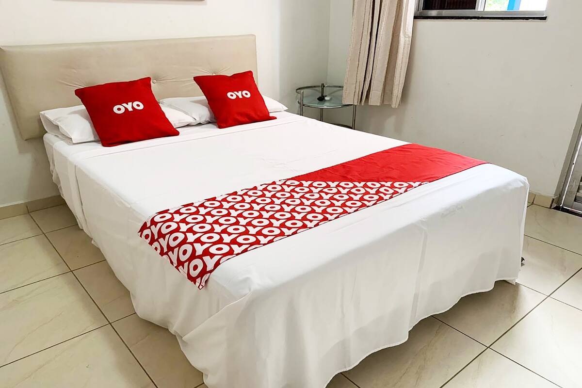 A bed with a white and red bedspread, two red pillows with 'OYO' text, a small bedside table, and a window with curtains.