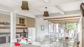 A modern kitchen with white cabinets, a microwave, and a refrigerator. There are white chairs and tables arranged for dining. - OYO Praia Hotel Recanto do Tomé - Salvador (Salvador)