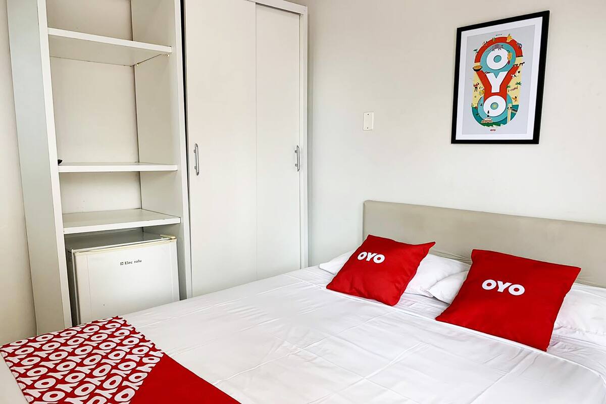A hotel room with a bed, two red pillows with the word "OYO" on them, a white headboard, and a framed picture on the wall.