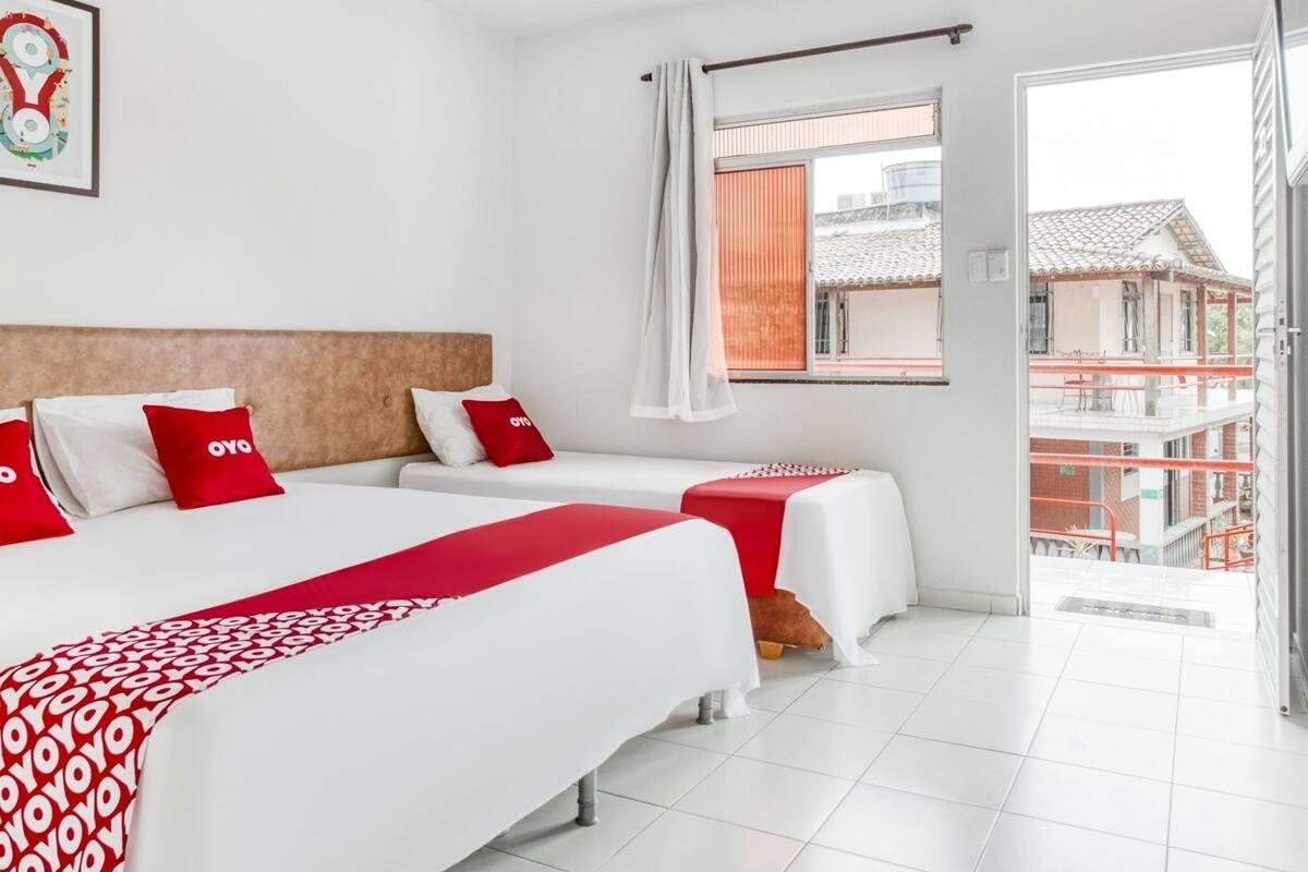 A hotel room with two beds, a window with a view of buildings, and a balcony.