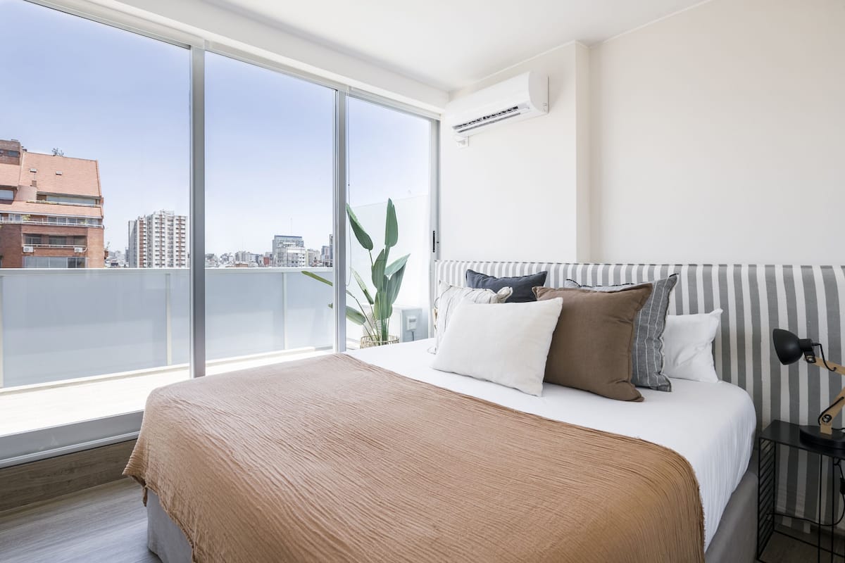 A modern hotel room with a large bed, a view of the city through floor-to-ceiling windows, and a small side table with a lamp.