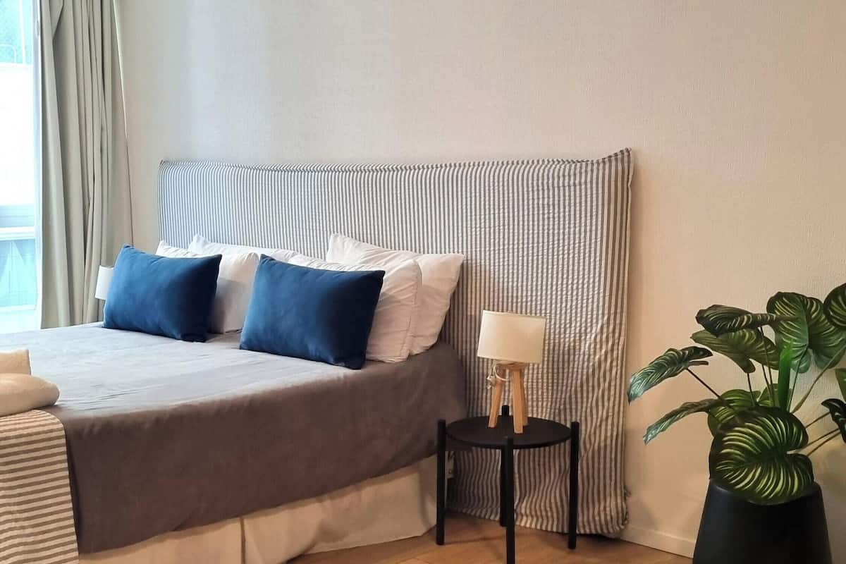 A hotel room with a bed, a bedside table with a lamp, a potted plant, and a view of the outside.