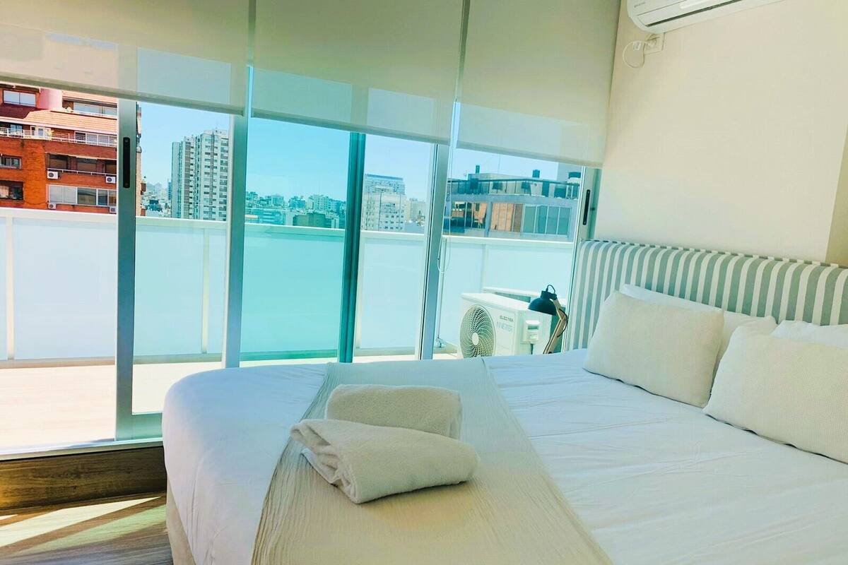 A hotel room with a large bed, a view of the city through floor-to-ceiling windows, and a balcony with a view of the water.