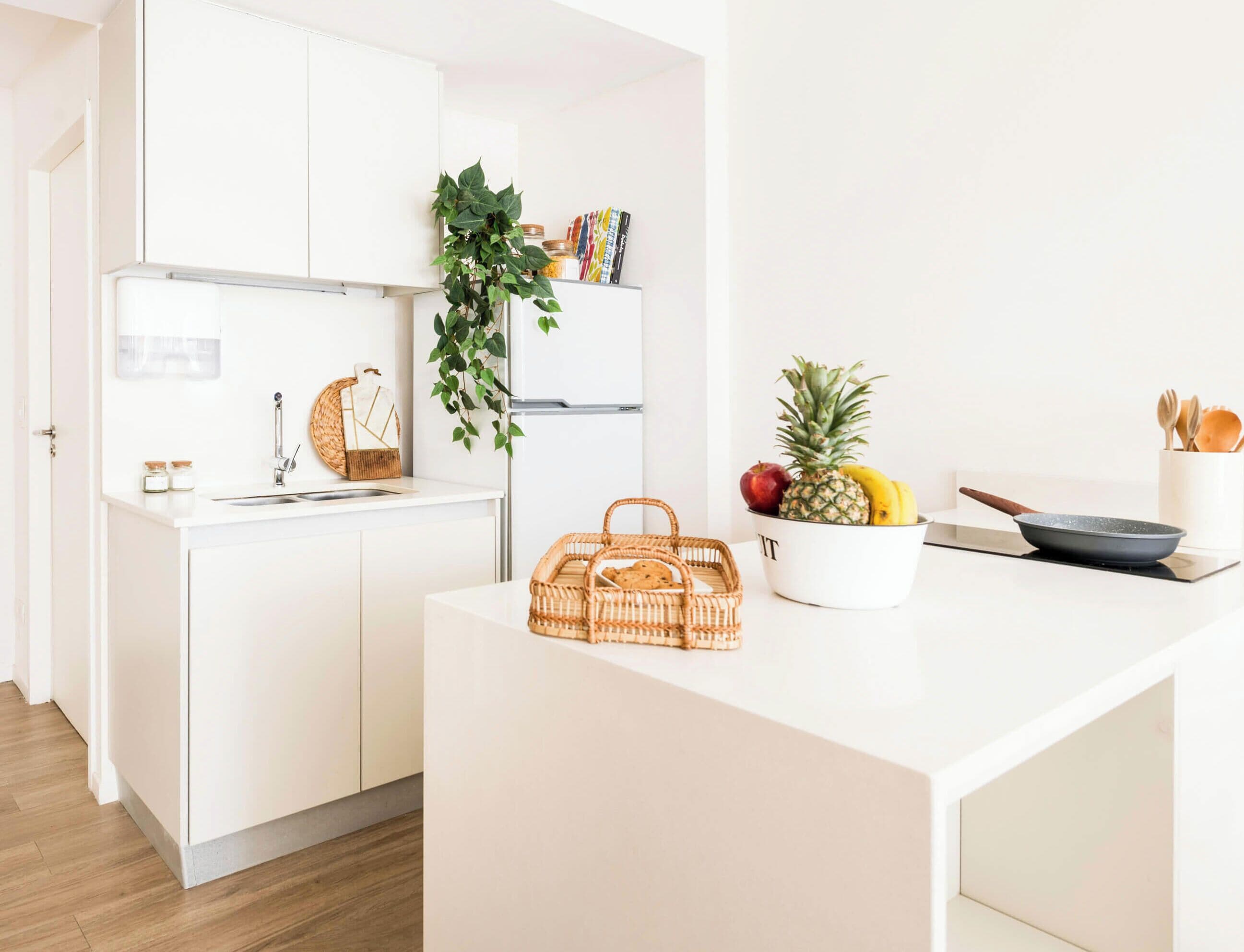 A modern kitchen with white cabinetry, a wooden floor, a hanging plant, a fruit bowl, and a wicker basket.