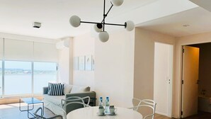 Una sala moderna con una mesa redonda blanca, sillas blancas, un sofá con almohadones a rayas y vistas a una masa de agua a través de amplias ventanas.