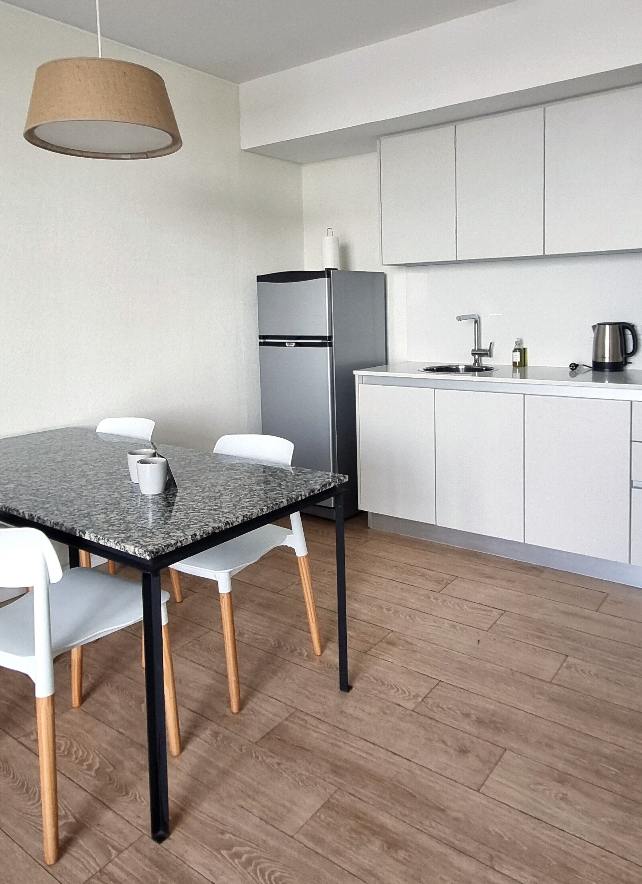 A modern kitchen with white cabinets, a stainless steel refrigerator, a wooden dining table with chairs, and a hanging light fixture.