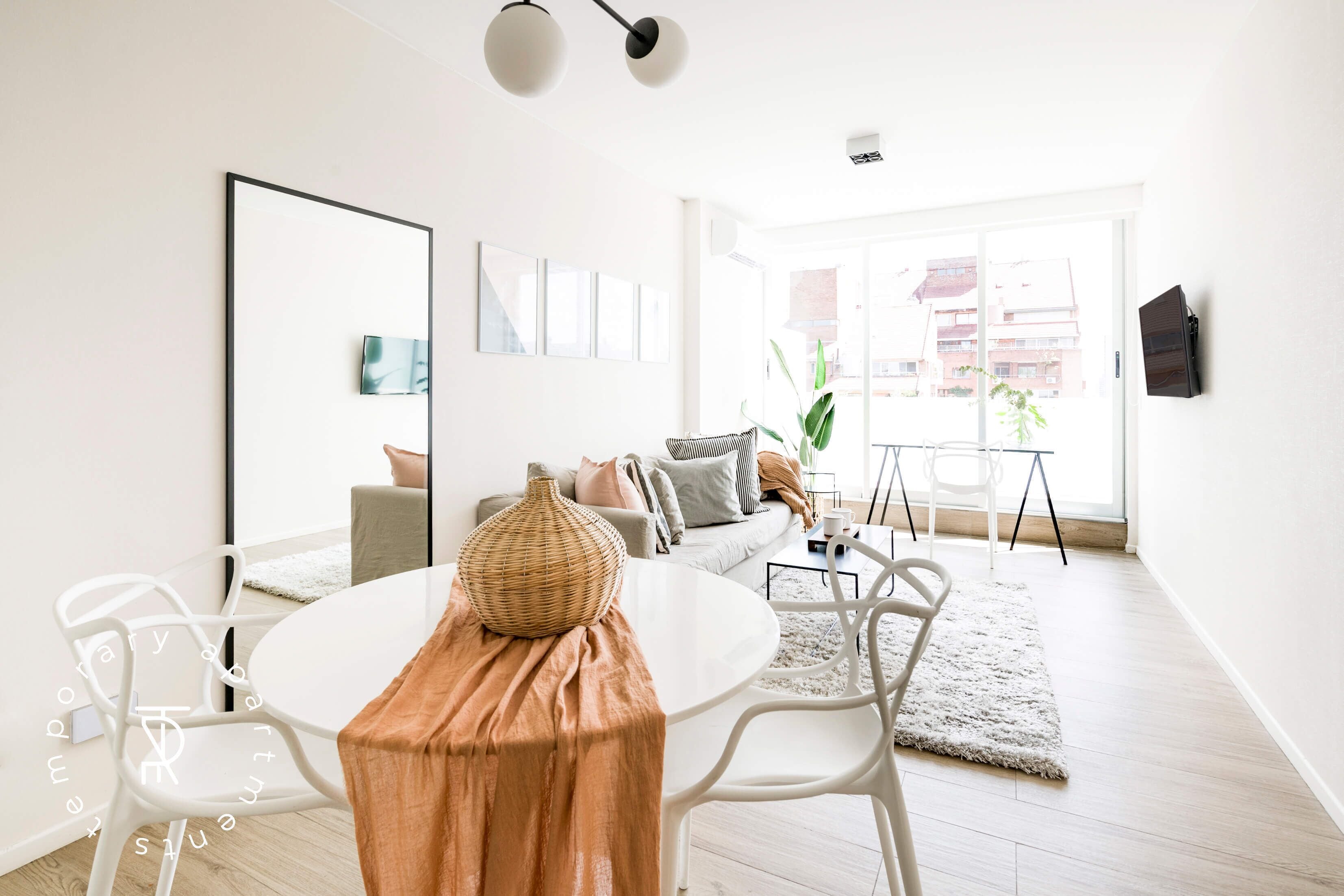 A modern living room with a white sofa, a dining table with chairs, and a large window with a view of buildings.