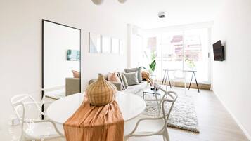 A modern living room with a white sofa, a dining table with chairs, and a large window with a view of buildings.