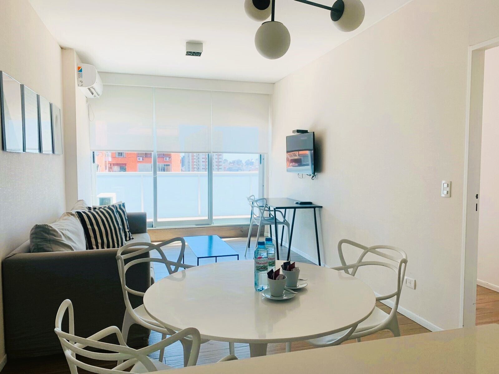 A modern living room with a round dining table, white chairs, a grey sofa with striped pillows, and a large window offering a city view.