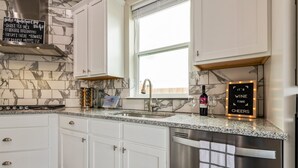 A modern kitchen with white cabinets, a granite countertop, a stainless steel dishwasher, and a window with a view. - Stay at Jake's Kalahari! Brand new house! (Round Rock)