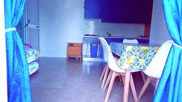 A dining area with a table and chairs, a kitchen in the background, and blue curtains framing the space. - Apartment nr. 2 in Villa Daniel, surrounded by greenery in VIESTE Gargano in P. (Vieste)