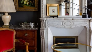 A room with a dark wall, a red chair, a white fireplace, and a mirror reflecting the room. - Moments From Monet, by Veeve (Paris)