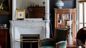 A room with a fireplace, a large mirror, a green armchair, a patterned rug, and a wooden cabinet with books. - Moments From Monet, by Veeve (Paris)