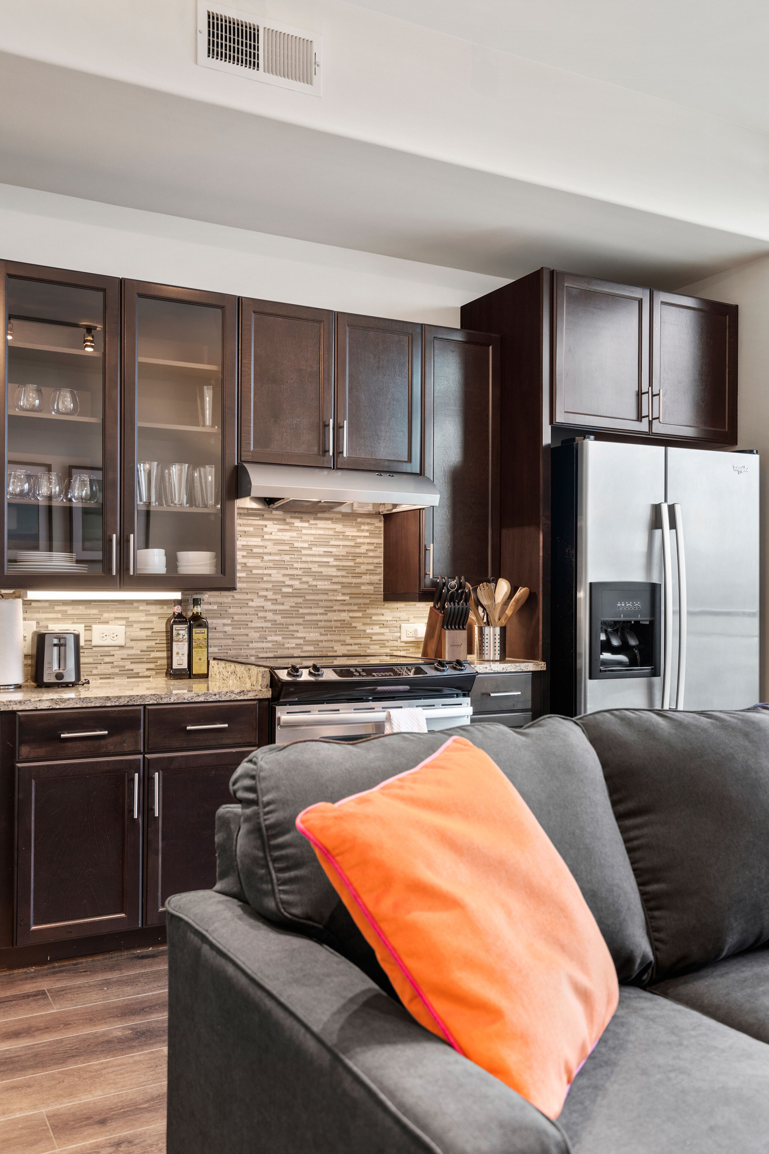 Una cocina moderna con armarios de madera oscura, electrodomésticos de acero inoxidable y un sofá gris con un cojín naranja.