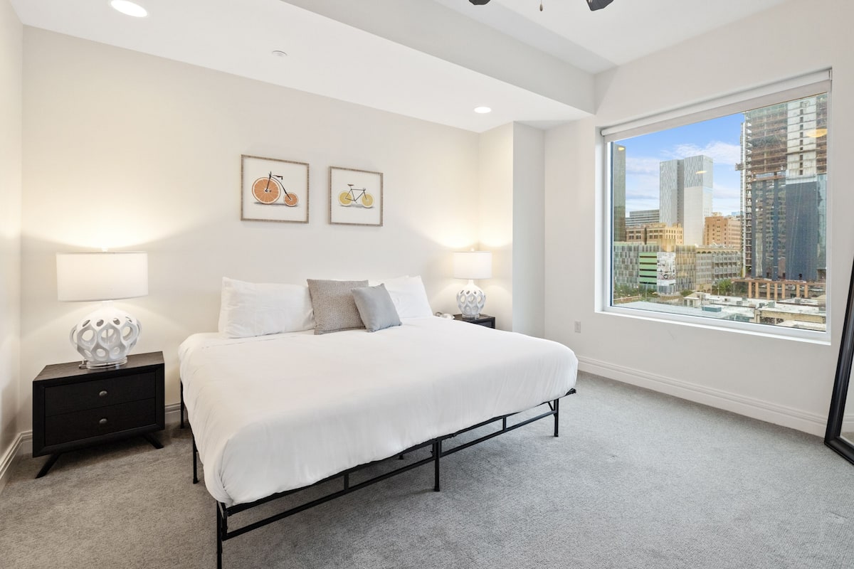 A modern bedroom with a large bed, two bedside tables with lamps, and a city view through a large window.