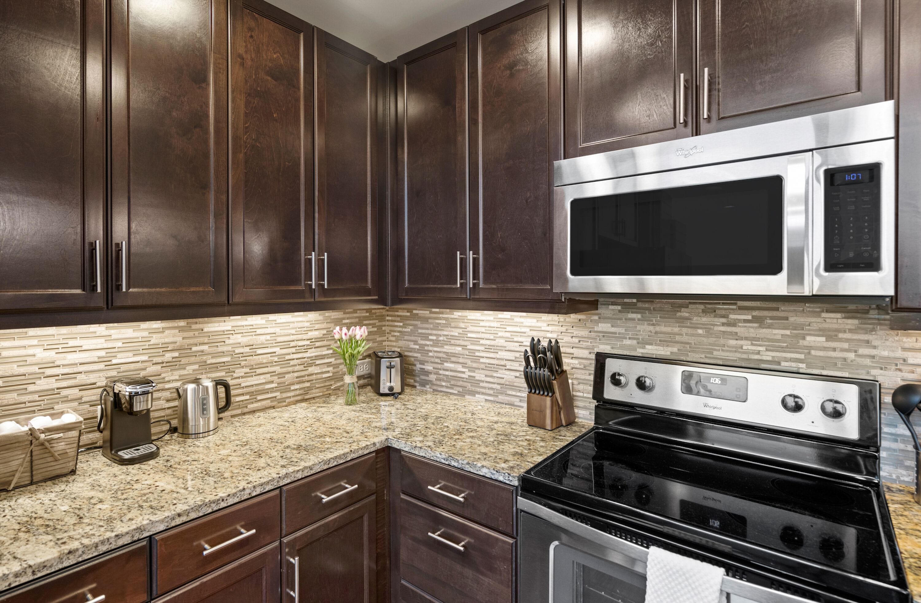 A kitchen with dark wood cabinets, a stainless steel microwave, a black stove, a granite countertop, and a tiled backsplash.