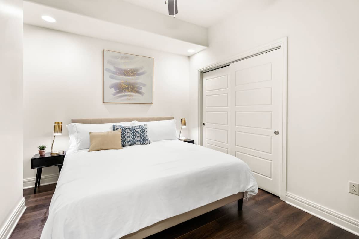 A bedroom with a large bed, a nightstand with a lamp, a wall-mounted abstract painting, and a white door.