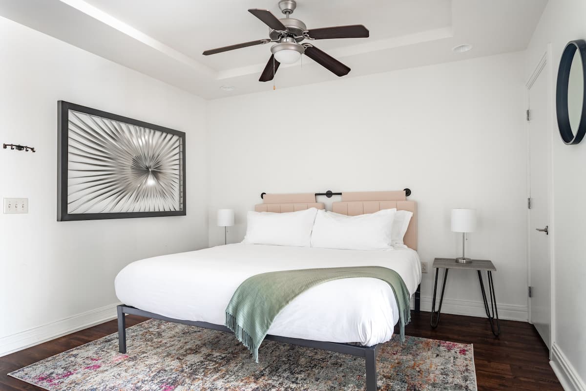 A bedroom with a bed, a ceiling fan, a framed abstract artwork, a bedside table with a lamp, and a door.