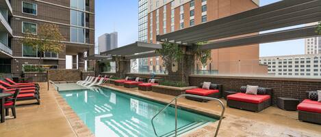 A rooftop pool area with red and black lounge chairs, a pool, and a view of a cityscape.