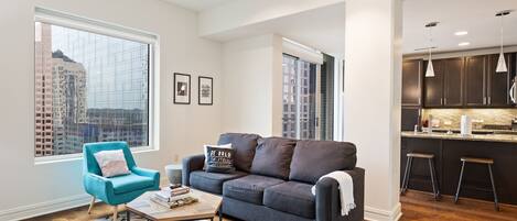 A modern living room with a sofa, a teal armchair, and a coffee table. There is a large window with a city view.