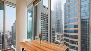 Una habitación de hotel moderna con un amplio ventanal que ofrece vistas a la ciudad, una mesa de comedor de madera y sillas blancas.