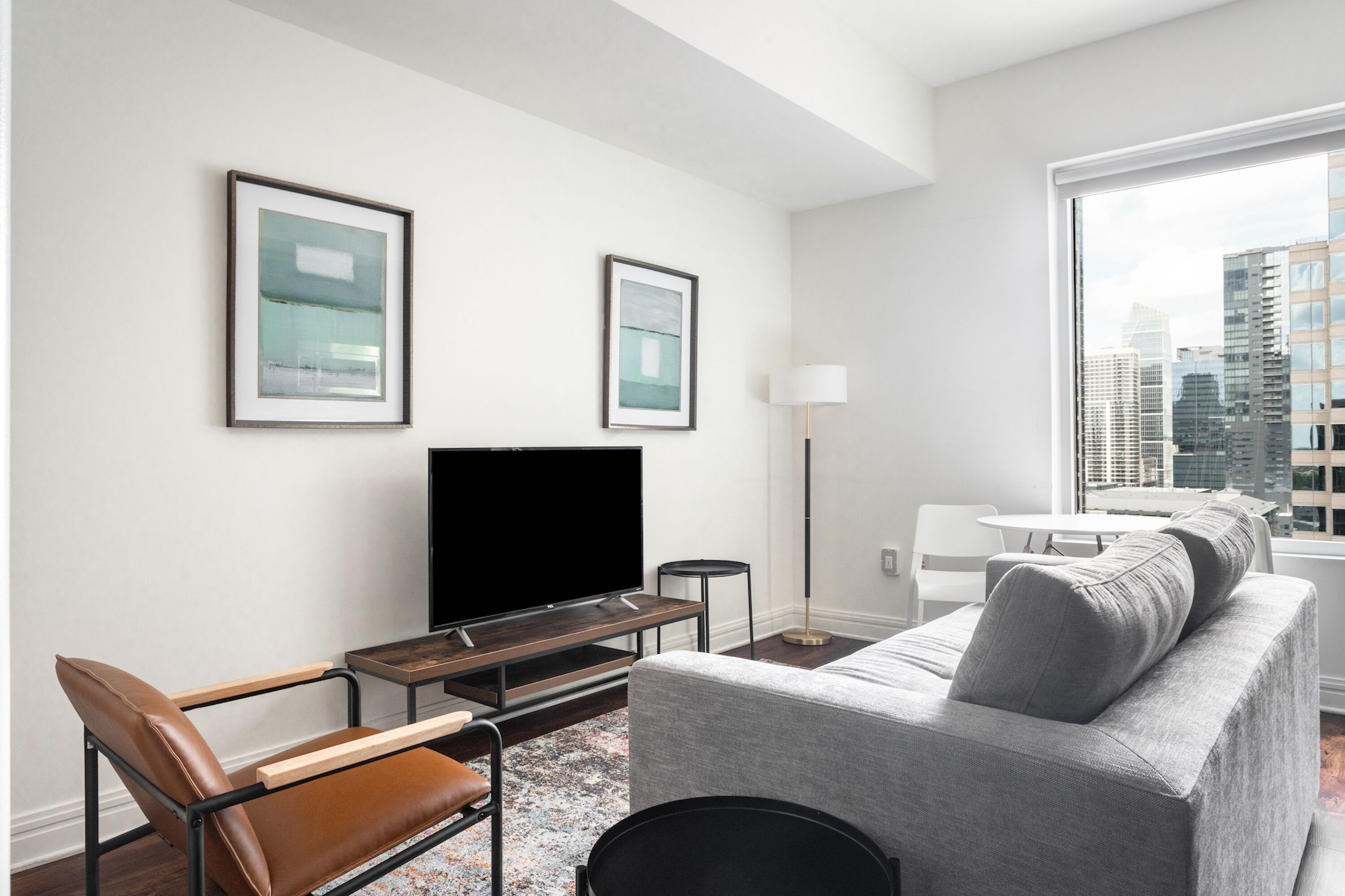 A modern living room with a flat-screen TV, a grey sofa, a brown leather chair, and a coffee table. There are two framed pictures on the wall and a floor lamp.