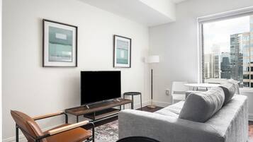 A modern living room with a flat-screen TV, a grey sofa, a brown leather chair, and a coffee table. There are two framed pictures on the wall and a floor lamp.
