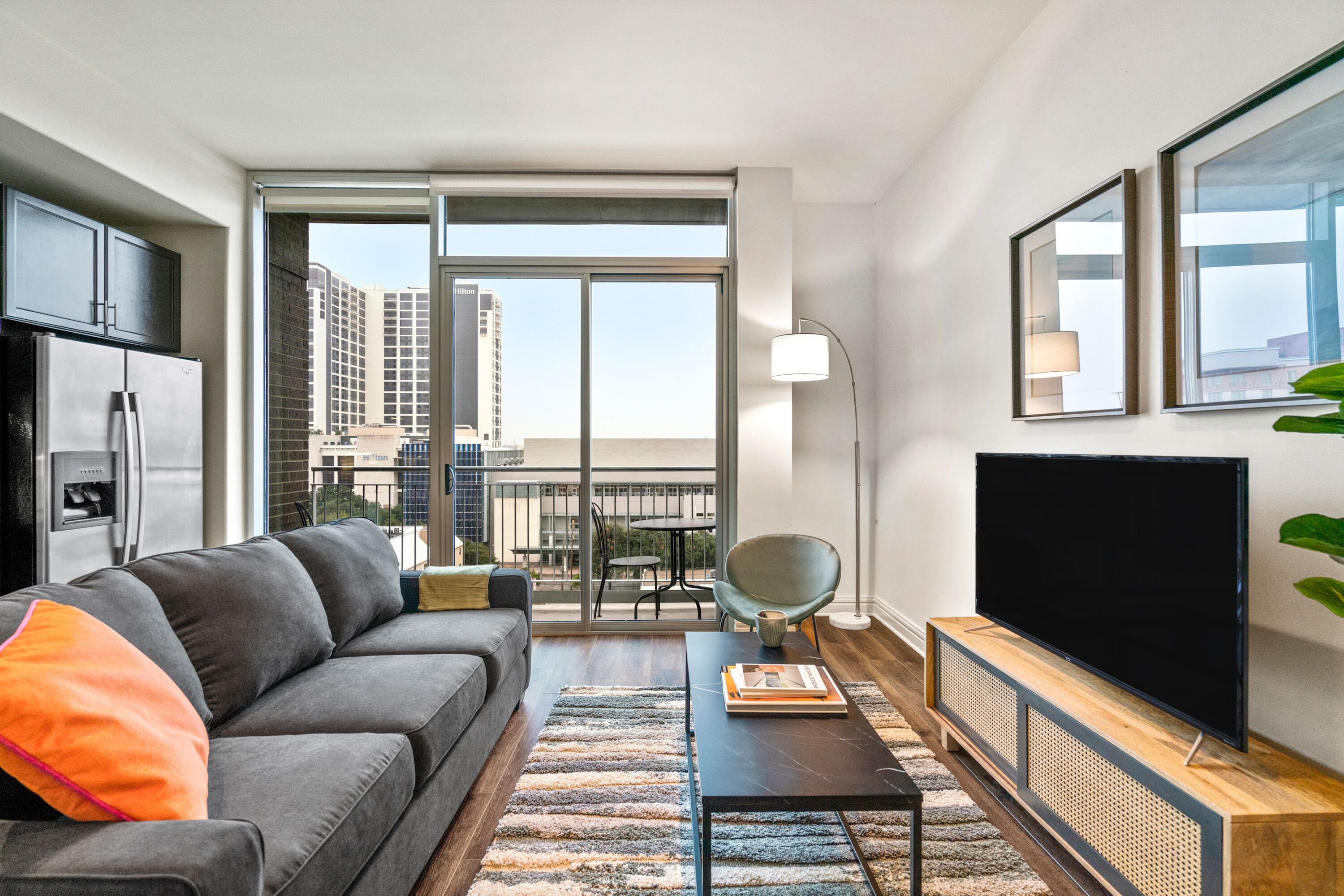 A modern living room with a grey sofa, a flat-screen TV, a coffee table, and a balcony with a view of the city.