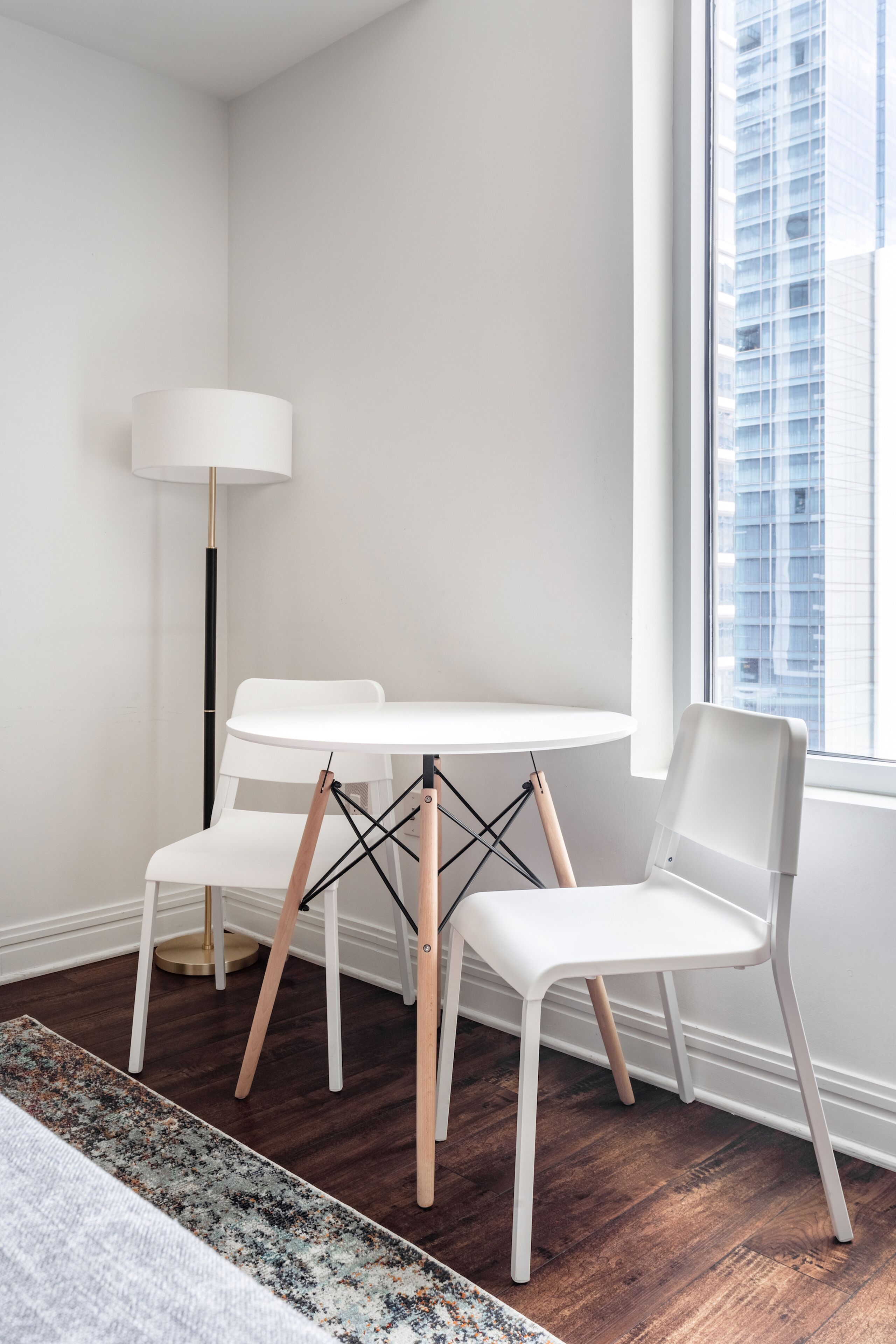 A modern room with a white table and chairs, a floor lamp, and a view of a building through a window.