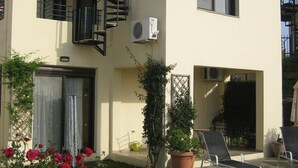 Een modern, twee verdiepingen hoog gebouw met een balkon, een kleine tuin met potplanten en buiten zitplaatsen.