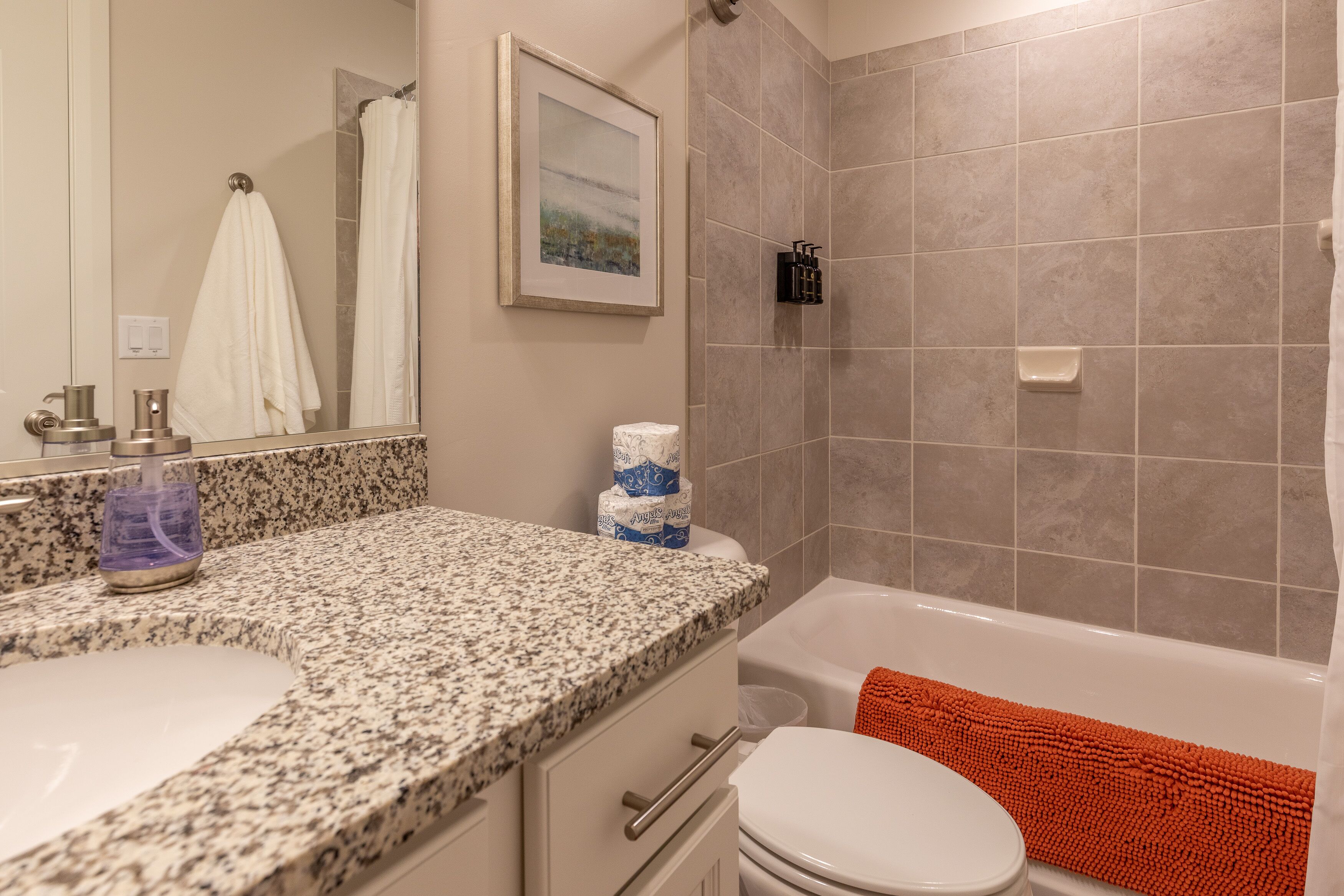 Ein Badezimmer mit einer Granitplatte, einer Toilette, einem Waschbecken, einer Dusche mit beige fliesenverkleideter Wand und einem gerahmten Bild an der Wand.