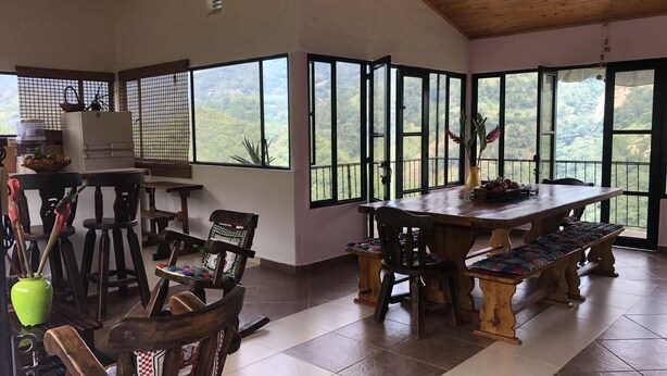 Un coin repas avec une table et des chaises en bois, de grandes fenêtres donnant sur un paysage verdoyant et un ventilateur de plafond.