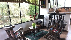 Une pièce chaleureuse avec un fauteuil à bascule, une petite table sur laquelle est posé un vase, et une vue sur la verdure à l’extérieur.